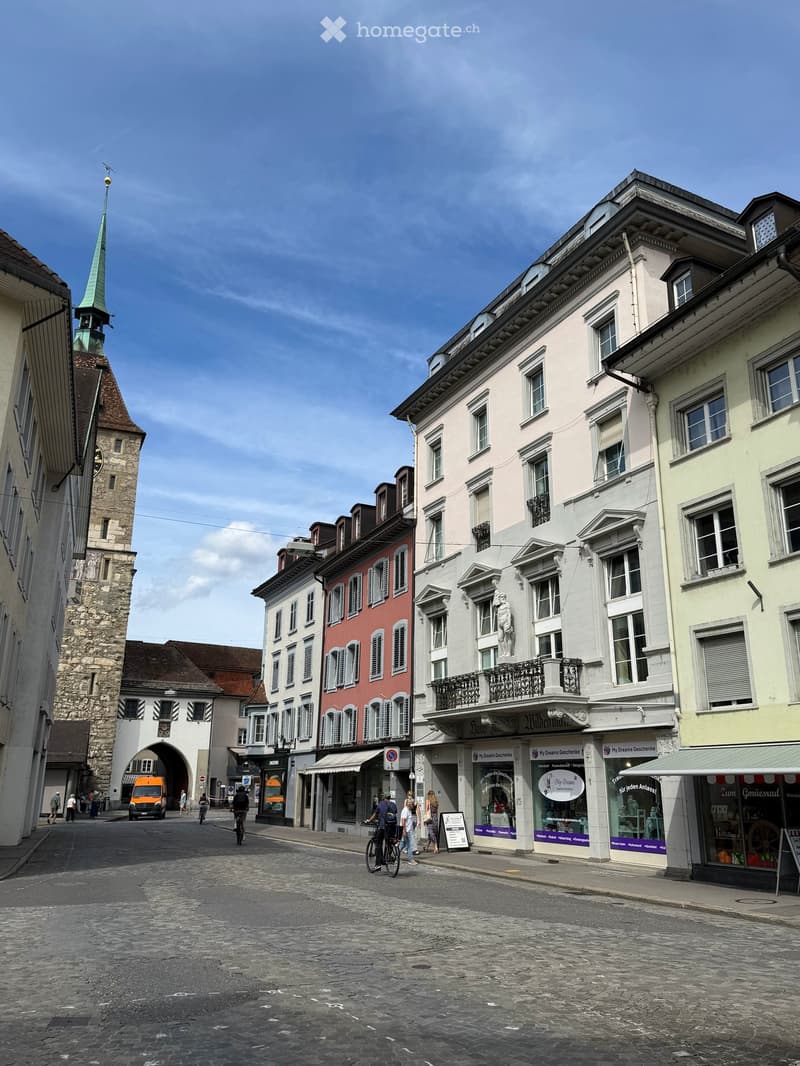 A historic European town square with multi-story buildings, a church tower, and pedestrians. The street is paved with cobblestones, and storefronts line the sidewalk.