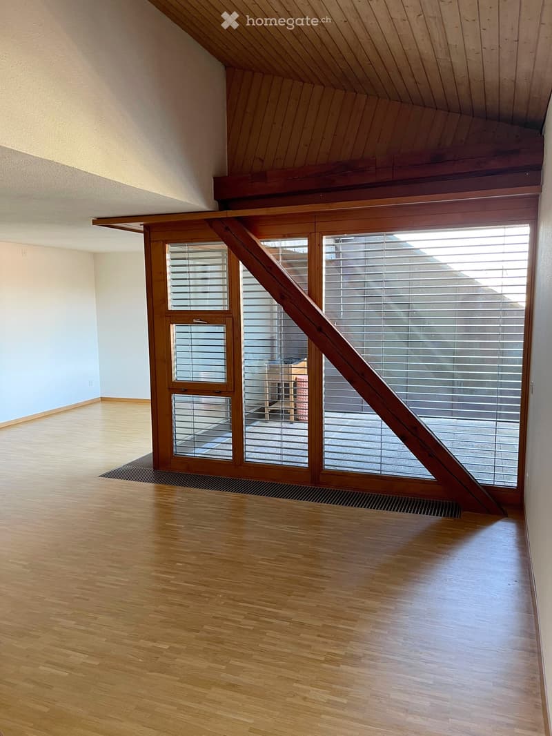 Bright, empty room with wooden flooring and large windows with blinds. The ceiling has exposed wooden beams, and the walls are painted in a light color.