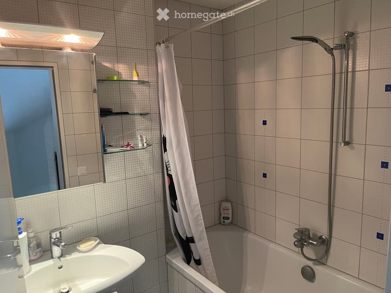 Modern bathroom with a white tiled wall, a sink with a mirror cabinet, a bathtub with a showerhead, and a shower curtain. The mirror cabinet has shelves with various toiletries.