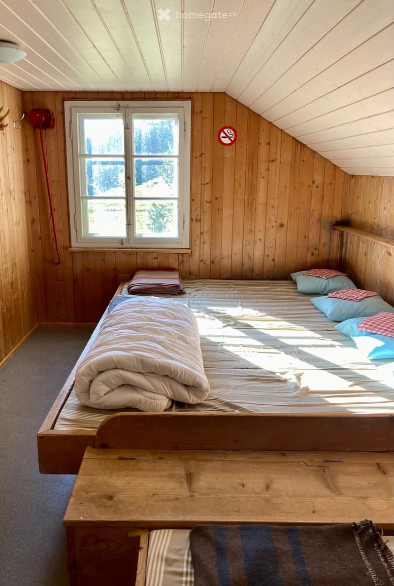 Cozy attic bedroom with wooden walls and slanted ceilings, featuring a single bed with pillows and a folded blanket, a wooden table, and a window offering natural light.