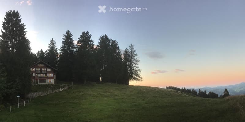 A two-story wooden house with white trim and a balcony, situated on a grassy hill surrounded by tall trees and a scenic mountain view at sunset.