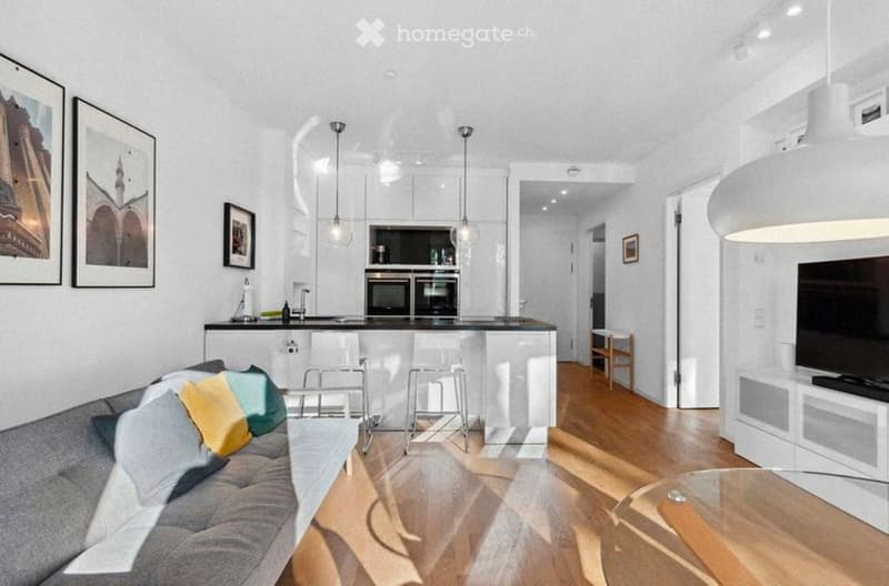 Open-plan living and dining area with a grey sofa, coffee table, bar stools, kitchen island, and modern appliances. The space features pendant lights, framed pictures, and a TV stand.