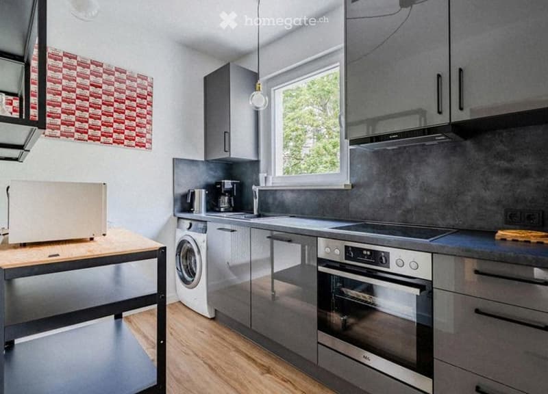 Modern kitchen with grey cabinets, stainless steel appliances, a wooden table, and a window overlooking greenery.