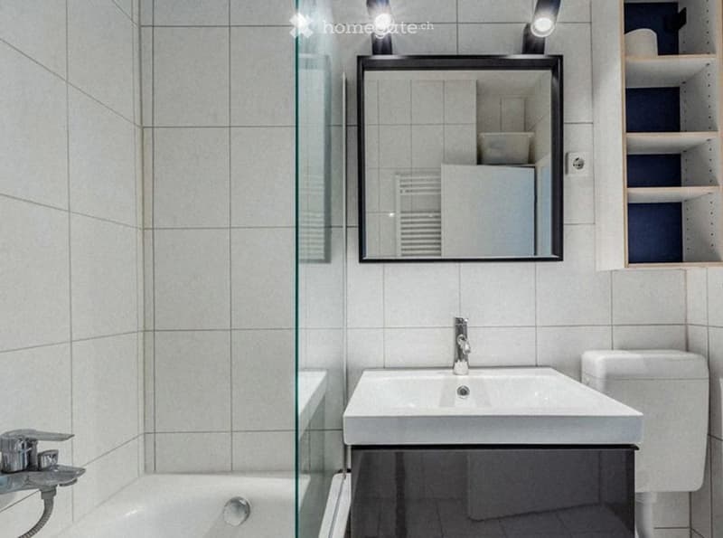 Modern bathroom with white tiles, a large mirror, a sink, a toilet, and a bathtub. The space includes a glass shower enclosure and a wooden shelf with storage items.