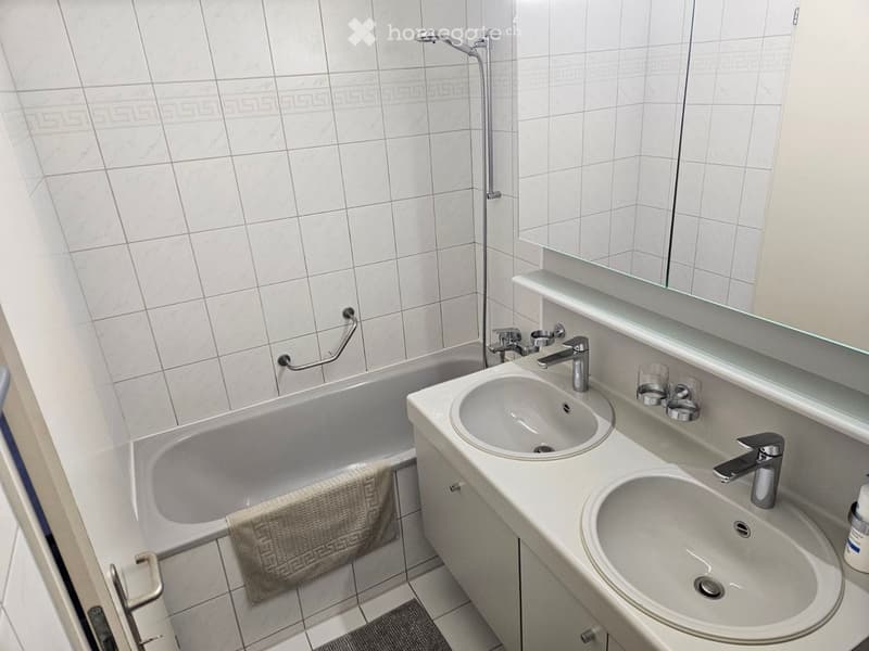 Modern bathroom with white tiles, a bathtub, double sink, and a large mirror above the sink.