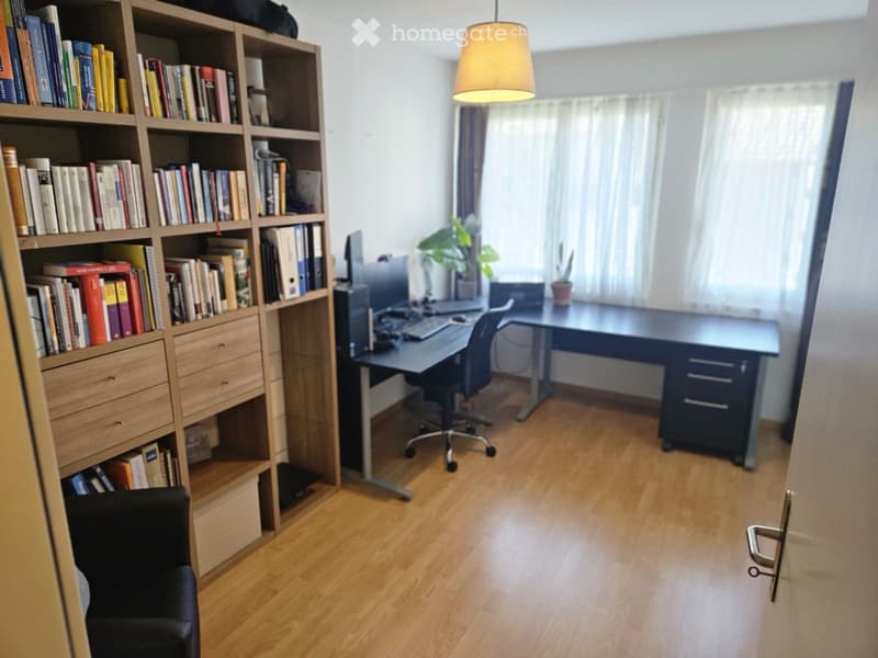 A well-lit study or home office with wooden flooring, a large bookshelf filled with books, a desk with a computer setup, and a comfortable chair.
