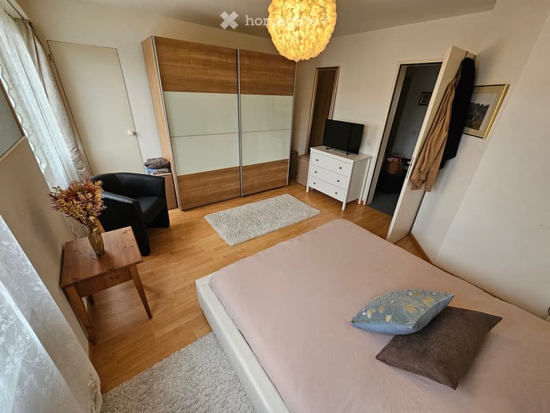 Cozy bedroom with a white bed, nightstand, wooden wardrobe, black armchair, and a small table with a vase of flowers. The room features wooden flooring and large windows with sheer curtains.