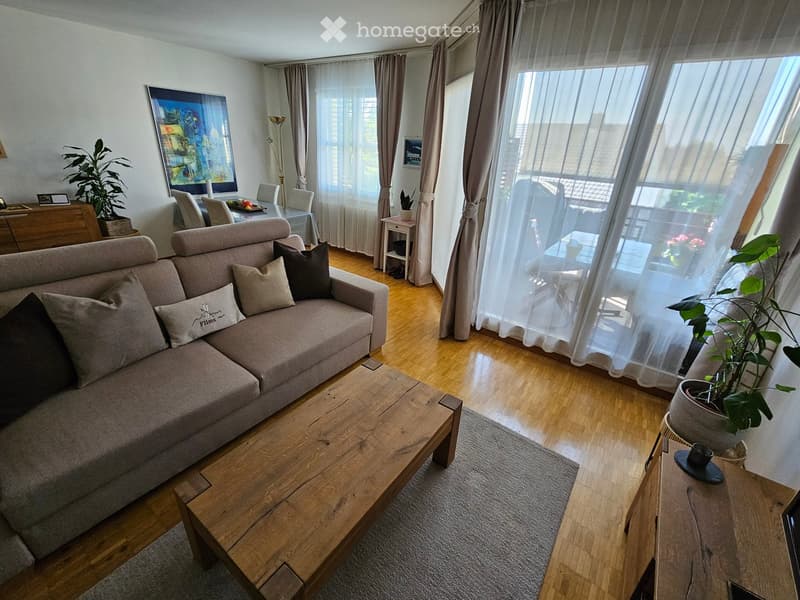 Modern living room with a grey sofa, wooden coffee table, side table, dining area with chairs, and large windows with sheer curtains.