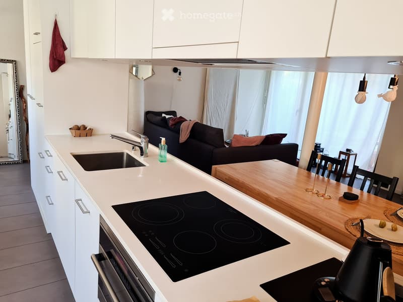 Modern kitchen with white cabinets, a central island, induction stove, sink, and a dining area with a wooden table and chairs. A sofa and mirror are visible in the adjacent living space.