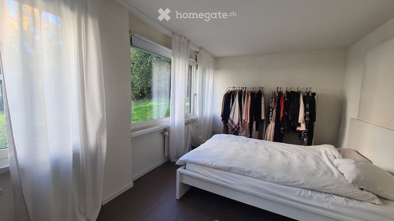 A tidy bedroom with a white bed, neatly made with pillows and a duvet, and a clothing rack with various garments hanging on it. The room has large windows with white curtains, allowing natural light to fill the space.