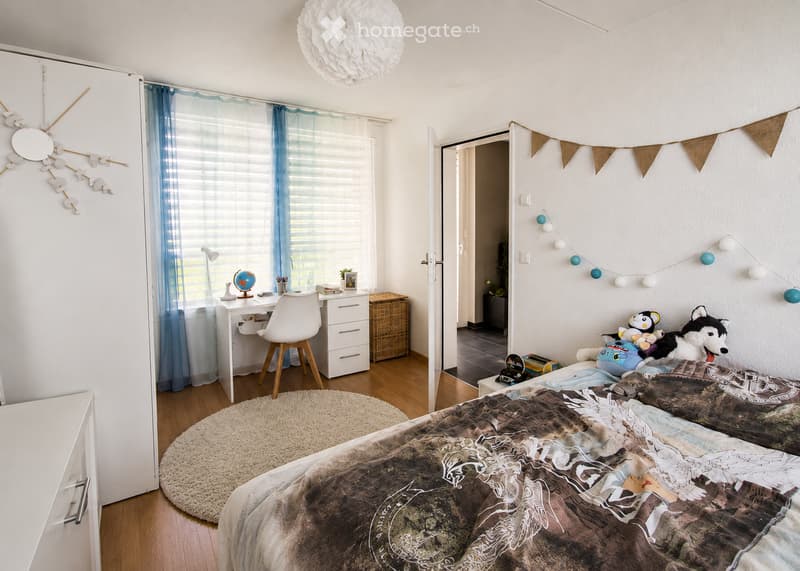 A well-lit bedroom with a large bed, a round shaggy rug, a white desk with a chair, a wardrobe, and decorative string lights. The room features a globe on the desk and a wicker basket near the window.