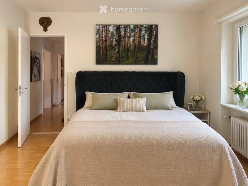 A well-lit bedroom with a large bed featuring a dark headboard and multiple pillows, a nightstand with a vase of white flowers, and a large forest-themed picture on the wall. The room has wooden flooring and an open doorway leading to another room.