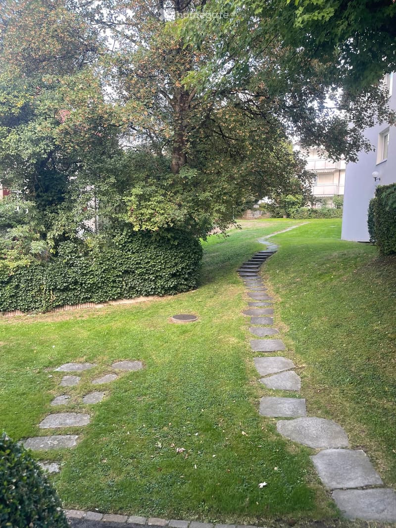 A well-maintained garden with a stone pathway leading to a grassy area, surrounded by trees and shrubs.