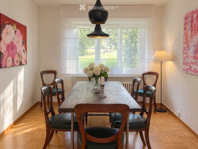 Dining room with wooden table, six chairs, vase with white flowers, candle, floor lamp, and two paintings on the walls.
