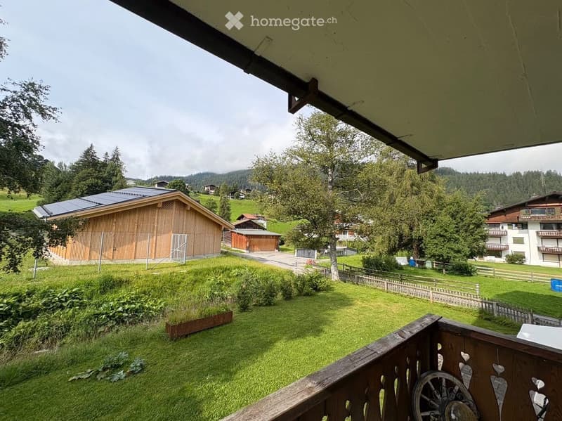 Balcony overlooking a green lawn, wooden fence, and distant houses with trees and mountains in the background.