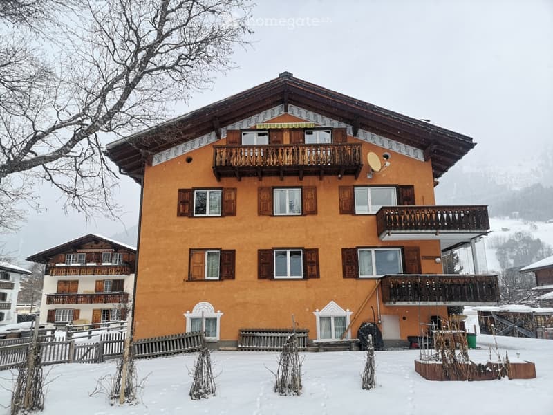 A two-story house with a yellow facade, wooden balconies, and traditional architectural details, surrounded by snow-covered ground and sparse vegetation.