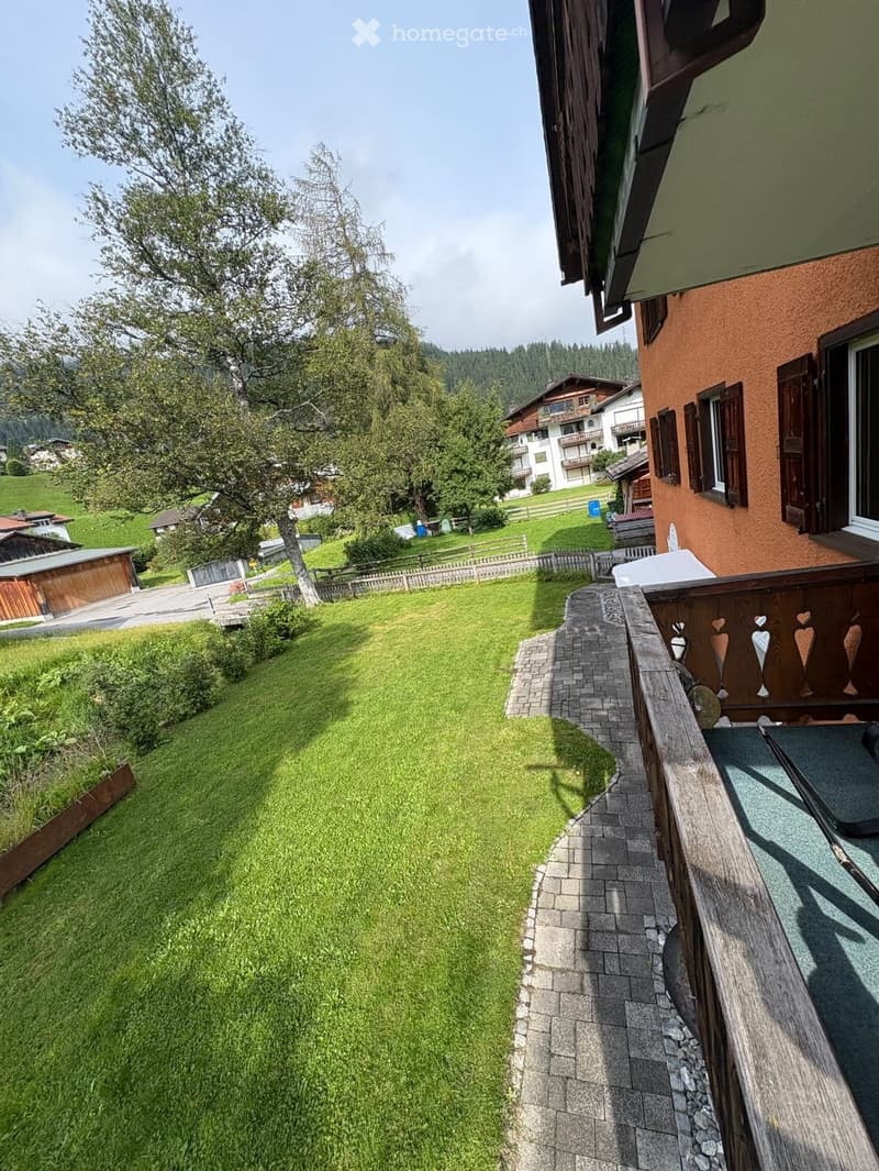 A well-maintained garden with a grassy lawn, a wooden fence, and a pathway leading to a wooden balcony with decorative elements.