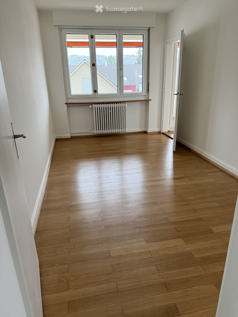 Empty room with wooden flooring, white walls, a radiator, and a large window with a view of neighboring rooftops.