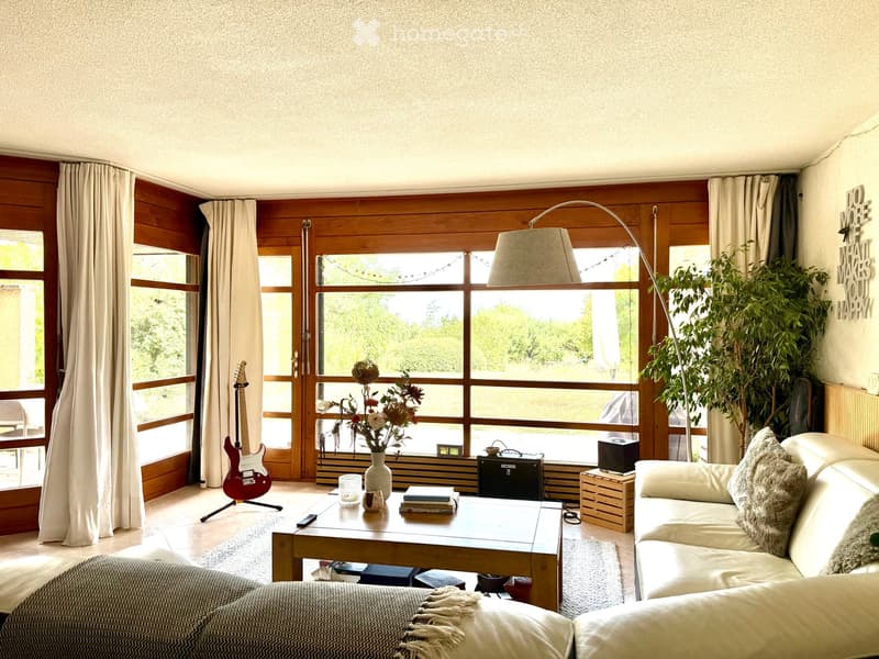Bright living room with large windows, wooden accents, a white sofa, coffee table, guitar, floor lamp, and decorative plants.