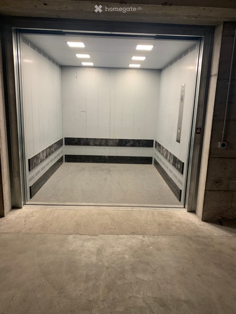 Empty concrete floor area with an open elevator shaft visible in the background, featuring white walls and ceiling lights.