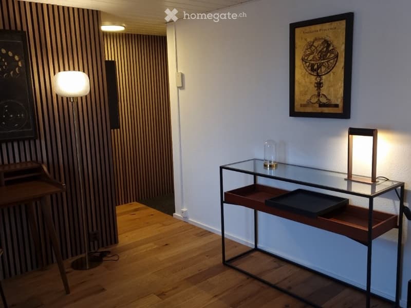 A modern, minimalist room with wooden flooring, a glass table with a wooden base, a floor lamp, a wooden stool, and framed artwork on the walls.