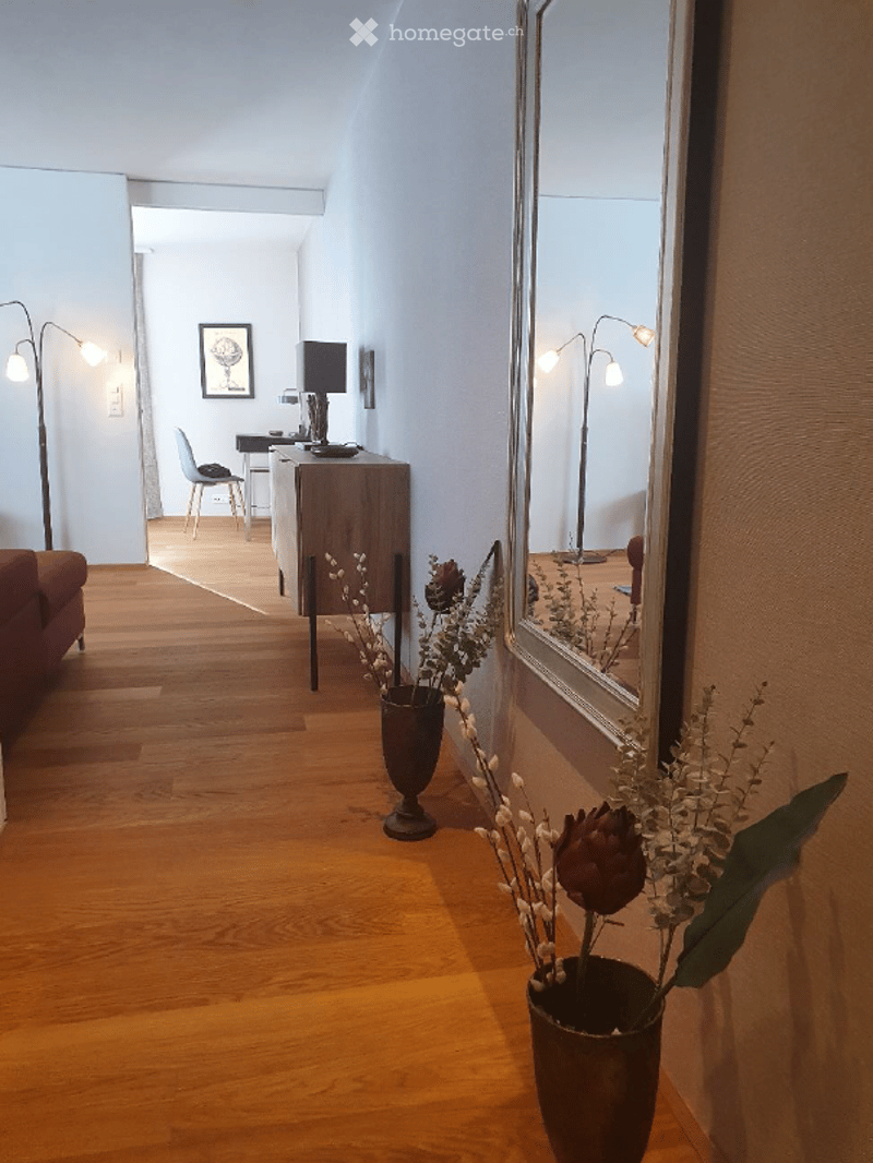 A well-lit hallway with wooden flooring, a large mirror on one wall, a vase with dried flowers, a red sofa on the left, and a small office area with a desk, chair, and lamp in the background.