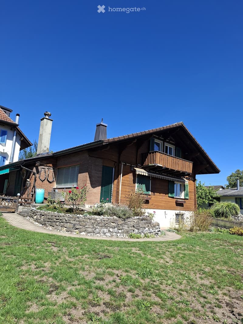 Two-story house with a wooden balcony, brick and wooden exterior, stone pathway, and garden area.