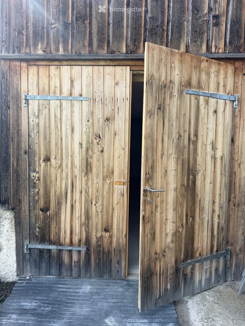 Wooden double doors with metal hinges and handles, set into a wooden wall, leading to a dark interior space.