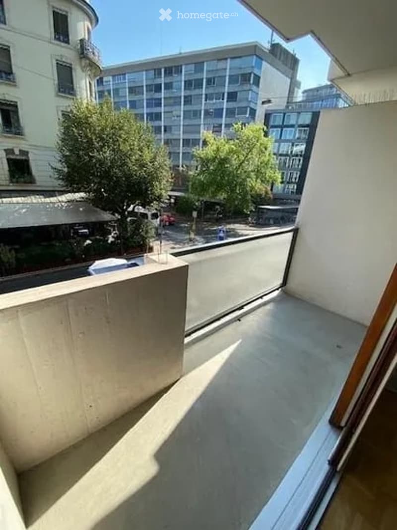 Balcony with a modern design, featuring a concrete floor and glass railing, overlooking a cityscape with trees and buildings.