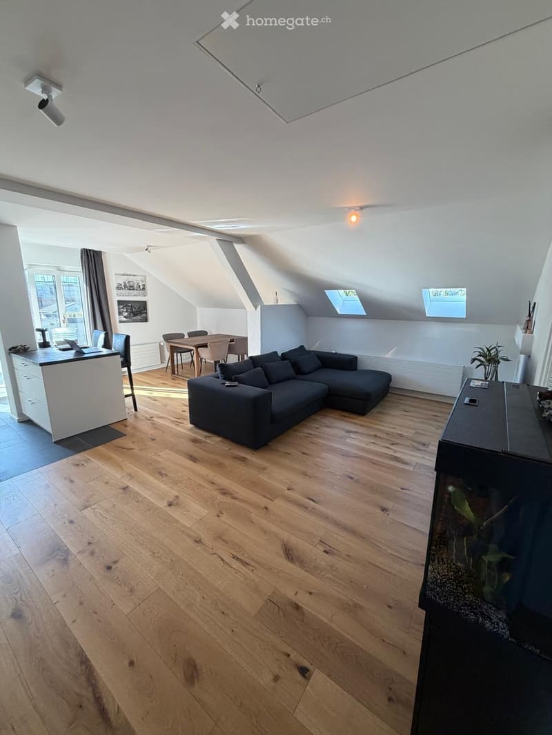 Modern living room with wooden flooring, a sectional sofa, dining area, and a fish tank. Features large windows and skylights.