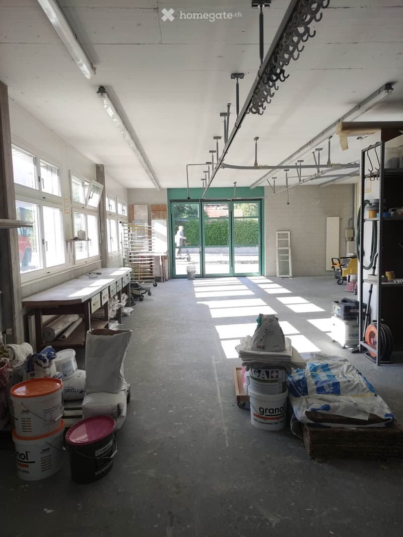Large, empty industrial-style room with concrete floors, white walls, and large windows. Contains storage shelves, buckets, and construction materials, but no furniture.