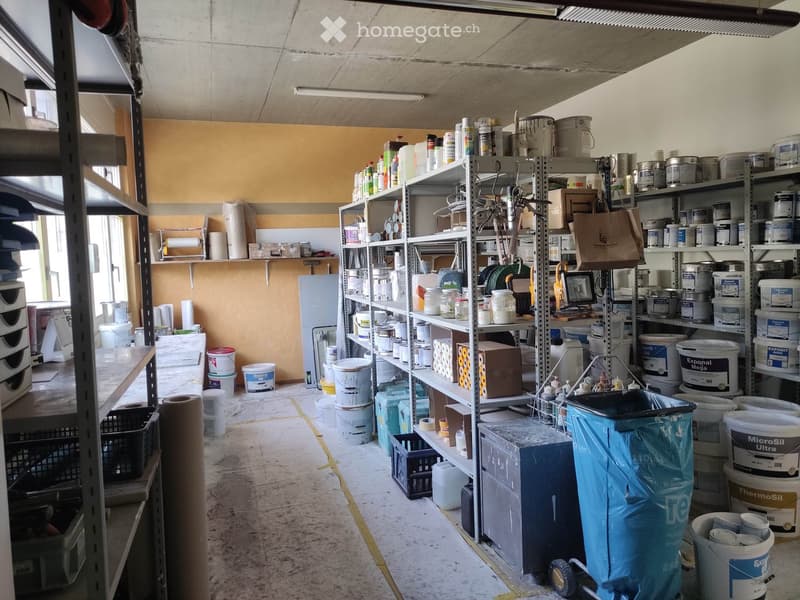 A storage area with metal shelving units filled with various paint cans, buckets, and tools. The space has a concrete floor and is well-lit with overhead lighting.