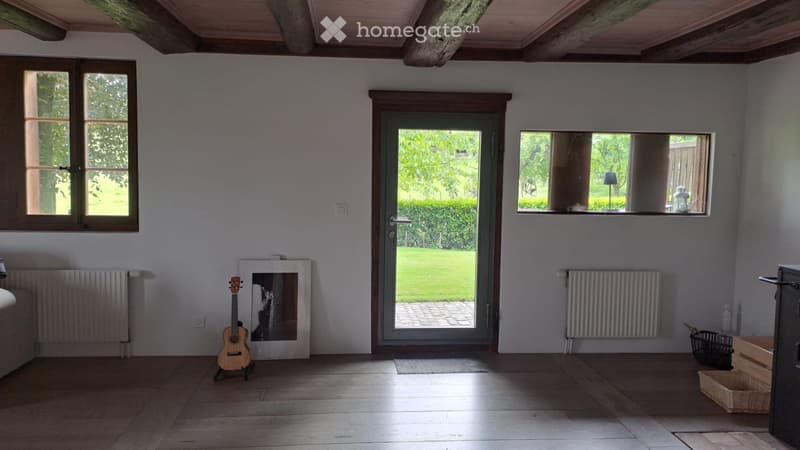 Bright, unfurnished room with wooden flooring, large windows, and a door leading to a garden. A guitar and a picture frame are placed on the floor.