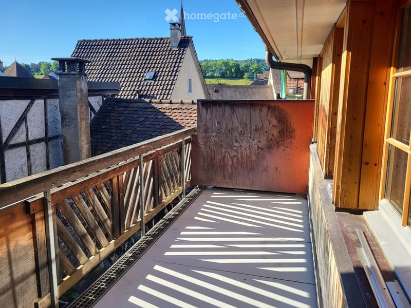Balcony with wooden railing, overlooking a traditional house with a tiled roof and chimney, surrounded by greenery.