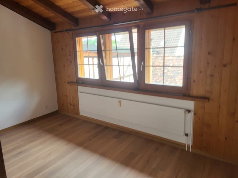 Empty room with wooden flooring, white walls, large window with wooden frame, radiator below the window, exposed wooden beams on the ceiling.