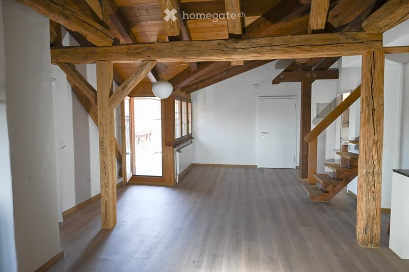Spacious hallway with wooden beams and flooring, leading to a staircase and an open doorway.