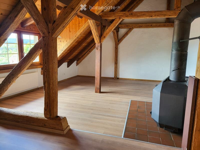 Empty attic space with wooden beams, wooden floor, and a large window offering a view of the outdoors. A wood-burning stove is present, but no furniture or furnishings are visible.