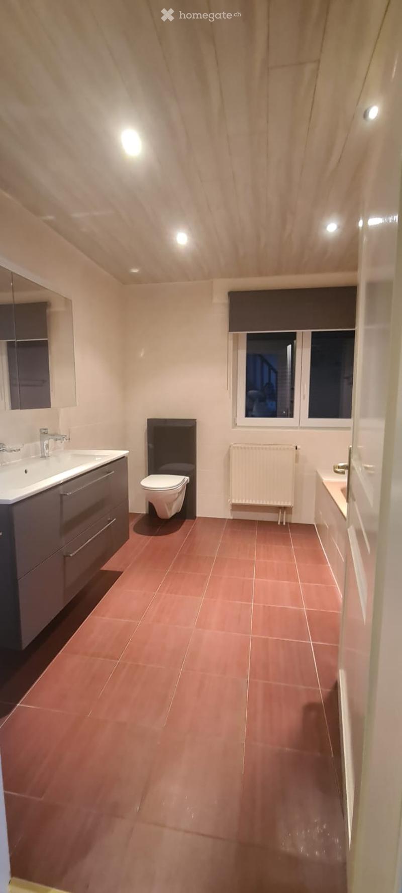 Modern bathroom with red tiled floor, white sink with faucet, toilet, radiator, and a bathtub. The room features a wooden slatted ceiling and a window with dark blinds.
