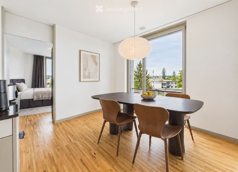 Modern open-plan living and dining area with wooden flooring, large windows offering a scenic view, a round dining table with four chairs, a bowl of fruit, a bed in the adjacent bedroom area, and a pendant light hanging from the ceiling.