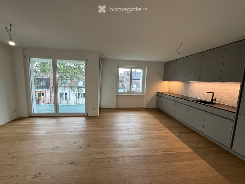 Empty room with wooden flooring, grey cabinets, and a sink, featuring large windows and a sliding glass door leading to a balcony.