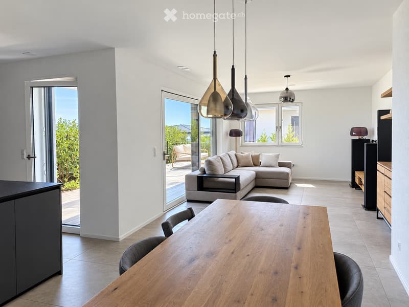 Modern living and dining area with a grey sectional sofa, wooden dining table, pendant lights, and large windows opening to a balcony.