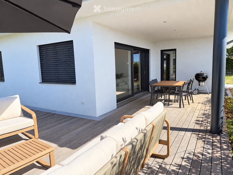 Modern outdoor patio with wooden decking, white walls, large sliding glass doors, outdoor furniture including a sofa, chairs, a table, and a grill.
