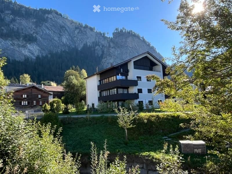 A modern multi-story building with balconies, surrounded by lush greenery and a scenic mountain backdrop.