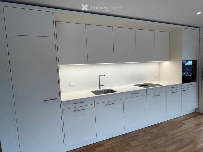 Modern kitchen with light grey cabinets, white countertops, a sink with a modern faucet, and a built-in stove. The space features sleek, contemporary design with integrated lighting above the counter.