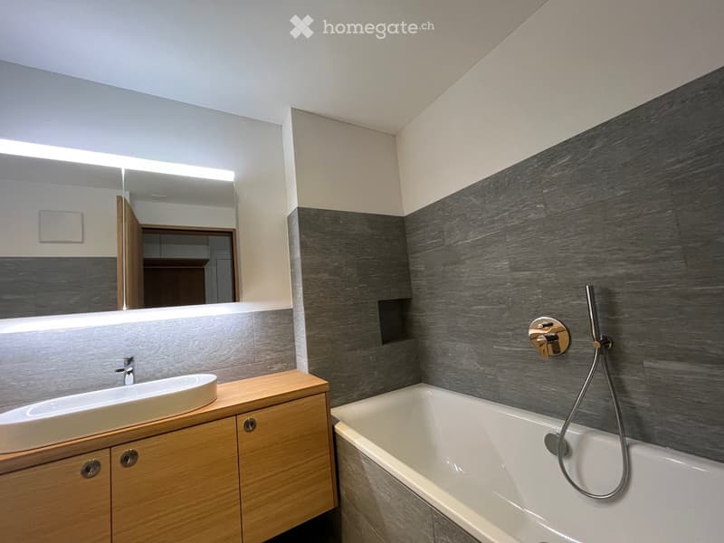 Modern bathroom with a wooden vanity, white sink, bathtub, and large mirror with built-in lighting. The walls feature a dark grey, wood-textured tile design.