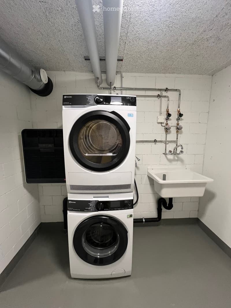 A compact laundry area with a stacked washing machine and dryer unit, a small sink, and exposed pipes.