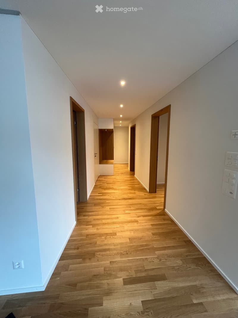 A clean, well-lit hallway with light wooden flooring and white walls. Features recessed ceiling lights, several doors, and electrical outlets on the walls.