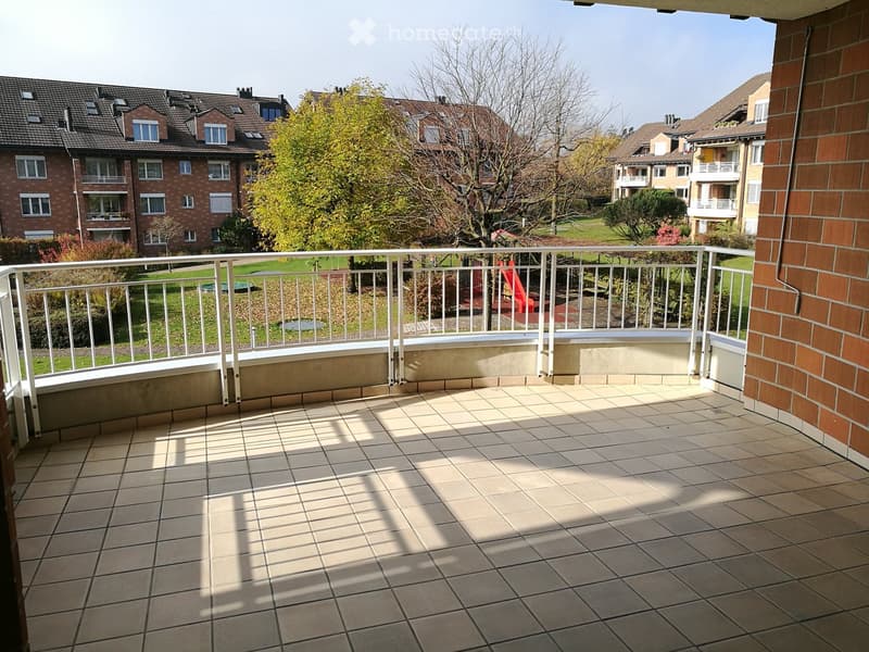 Balcony with tiled floor, white metal railing, overlooking a residential area with trees and buildings.