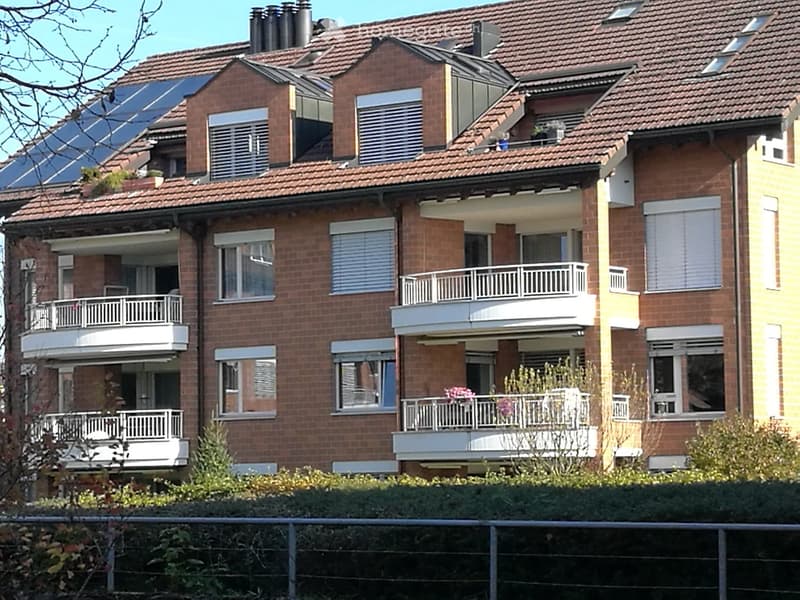 Multi-story brick apartment building with multiple balconies, solar panels on the roof, and surrounding greenery.