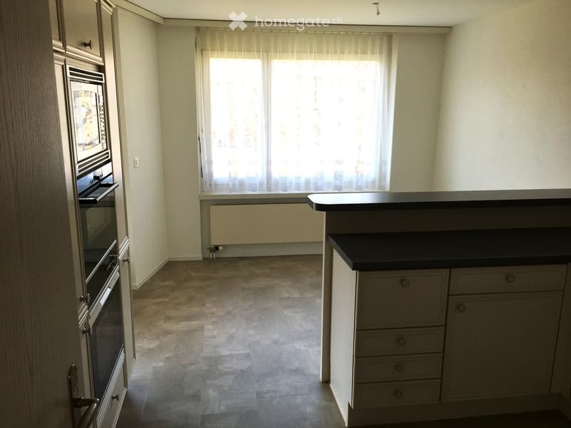 Empty kitchen with light-colored cabinets, a black countertop, and a built-in oven. The space features tiled flooring and large windows with sheer curtains.